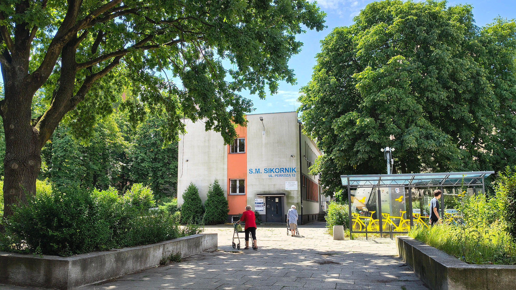 dwie kobiety z balkonikami spacerują na skwerze, obok wielkie drzewa i budynek spółdzielni