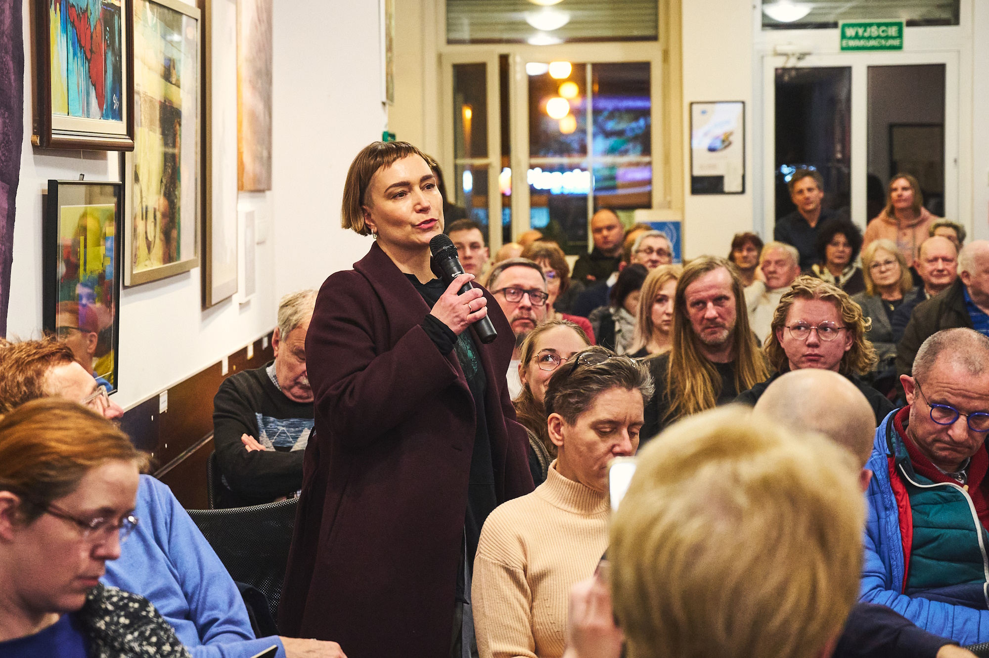 Kobieta mówi do mikrofonu, wokół niej siedzą ludzie
